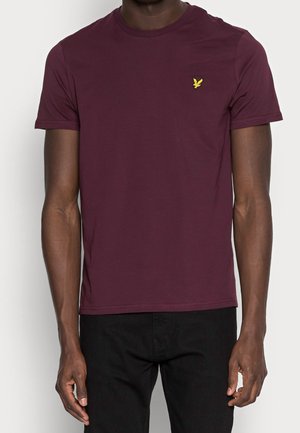 Mann trägt ein schlichtes burgunderfarbenes T-Shirt mit kurzen Ärmeln und einem kleinen gelben Vogel-Logo auf der linken Brustseite und schwarze Hose.