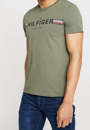 Mann trägt olivgrünes kurzärmeliges T-Shirt mit dem Schriftzug "Hilfiger New York" und Streifen, kombiniert mit dunkelblauen Jeans.