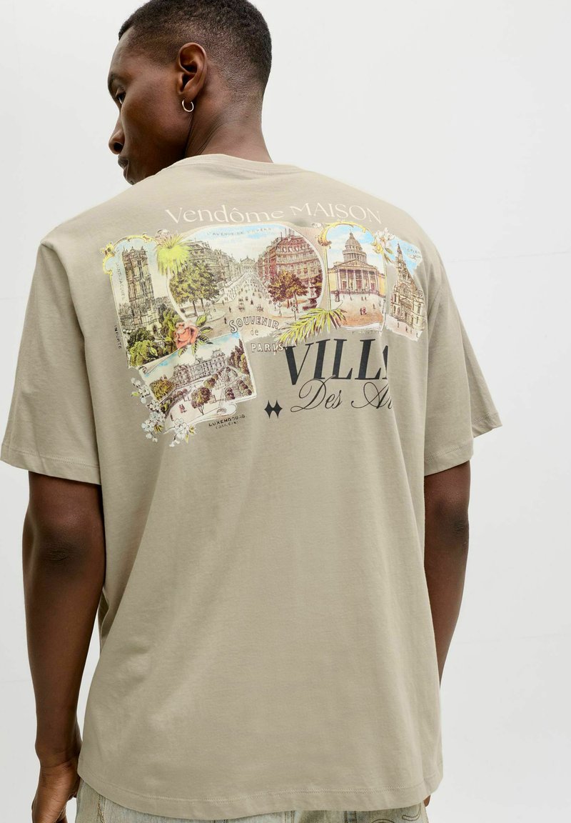 Mann trägt ein beige T-Shirt mit einem Vintage-Design von Pariser Sehenswürdigkeiten und den Texten „Vendôme Maison“ und „Villa Des Arts“ auf der Rückseite.