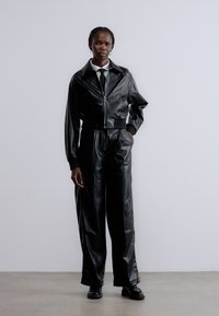 Svart kort skinnjacka med dragkedja framtill, kombinerad med lösa svarta skinnbyxor. Mjuk textur, minimalistisk design och skräddarsydd passform.