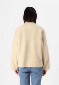 Beige fleecejakke med en lett teksturert overflate, dropped shoulders og en enkel krage. Kombinert med blå denimjeans.