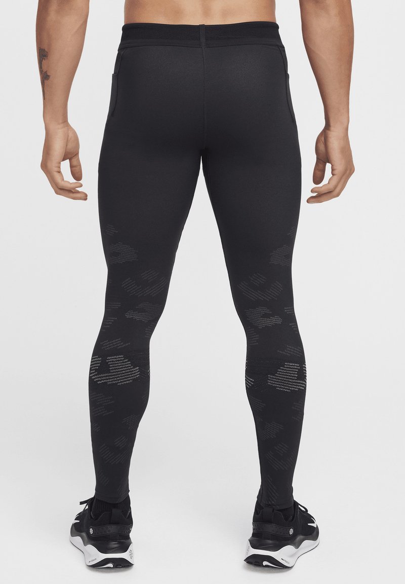 Homme de dos portant des leggings noirs à motif camouflage et des baskets noires sur un fond clair.