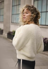 Fuzzy, hvid sweater med en afslappet pasform og faldne skuldre, båret over mørke bukser med hvide sidestriber, mod en sløret bybaggrund.