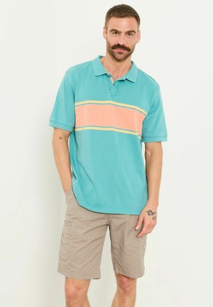 Mann mit Schnurrbart, der ein türkisfarbenes Poloshirt mit pfirsichfarbenem Streifen und beige Cargoshorts trägt, steht mit einer Hand in der Tasche.