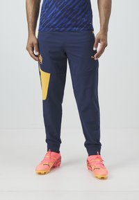 Marineblauwe sportbroek met een gele zijpaneel en logo, voorzien van een gestroomlijnd ontwerp en lichtgewicht stof. Gecombineerd met felroze sportschoenen.
