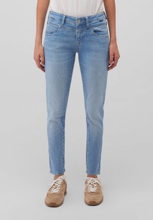 Frau in hellblauen Slim-Fit-Jeans und beigen Sneakers, die vor einem schlichten hellen Hintergrund steht.
