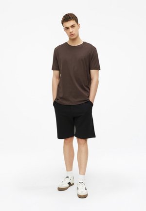 Junger Mann steht mit den Händen in den Taschen, trägt ein braunes T-Shirt, schwarze Shorts, weiße Socken und weiße Sneaker vor weißem Hintergrund.