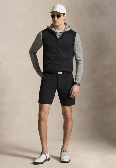 Άντρας με άσπρο καπέλο, γυαλιά ηλίου, γκρι φούτερ με κουκούλα, μαύρο γιλέκο με φερμουάρ, μαύρα σορτς, ρολόι και άσπρα παπούτσια που στέκεται μπροστά σε ουδέτερο φόντο.