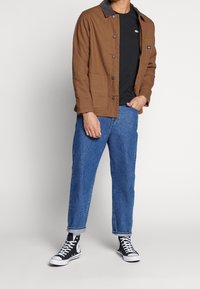 Brown canvas jacket with a dark collar, black t-shirt, light blue denim jeans, and black high-top sneakers with white accents.