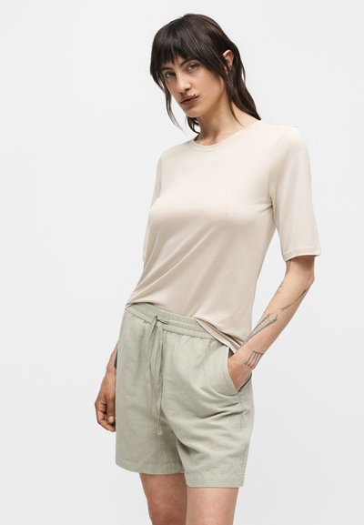 Γυναίκα με σκουρόχρωμα μαλλιά που φοράει μπεζ τοπ με κοντά μανίκια και ανοιχτό γκρι σορτς με κορδόνι, στέκεται με το ένα χέρι στην τσέπη σε λευκό φόντο.