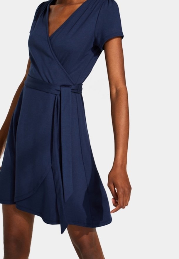 Femme portant une robe portefeuille bleu marine à manches courtes avec une ceinture nouée à la taille, debout devant un fond clair uni.