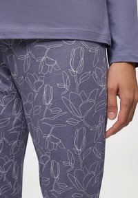 Top lungo viola a maniche lunghe con scollatura rotonda, abbinato a pantaloni viola con un motivo floreale bianco a contorno, tessuto dalla consistenza liscia.