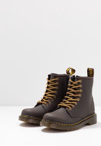 Brązowe skórzane buty za kostkę z żółtymi sznurówkami, bocznym zamkiem błyskawicznym oraz solidną czarną gumową podeszwą z żółtymi szwami.