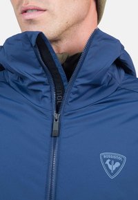 Navyblaue Jacke mit hohem Kragen, schwarzem Reißverschluss und Kapuze. Verfügt über ein hellblaues Rossignol-Logo auf der Brust und eine glatte Textur.
