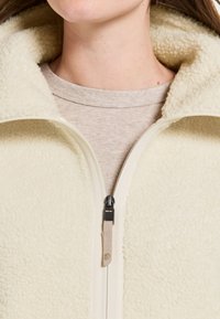 Crème-kleurige fleecejas met een zachte, gestructureerde oppervlakte en hoge kraag, voorzien van een voorrits in het zwart en beige details.