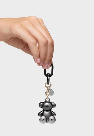 Ręka trzyma czarny metalowy karabińczyk z breloczkiem oraz małym czarnym pluszowym misiem i okrągłą metalową zawieszką.