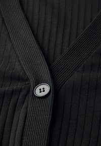 Schwarze gerippte Strickjacke mit tiefem V-Ausschnitt und einem runden schwarzen Knopf am Verschluss, die einen strukturierten Stoff hervorhebt.