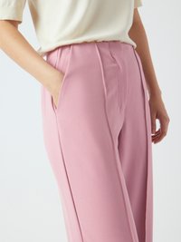 Persoon in hoge roze op maat gemaakte broeken met een hand in de zak en een kort mouw crèmekleurig top tegen een effen achtergrond.