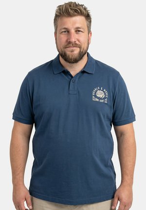 Bärtiger Mann mit kurzen Haaren, der ein blaues Polohemd mit dem Logo "Island Surf Co." trägt, steht vor einem schlichten hellen Hintergrund.