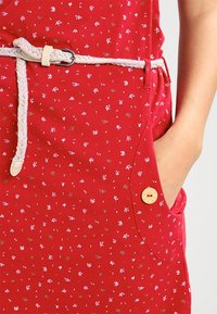 Punainen mekko, jossa on pieni valkoinen ja ruskea kukkakuosi, letitetty valkoinen vyö, edessä nappikiinnitteinen tasku, käsi taskussa.