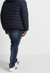 Persoon in een donkerblauwe gewatteerde jas, blauwe jeans en witte Adidas sneakers, staande tegen een effen witte achtergrond.