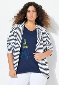 Stripet blazer i hvid og sort over en navyblå t-shirt med gul og blå tekst. Modellen bærer hvide bukser. Bløde materialer, afslappet design.