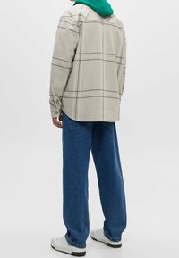 Beige geruite shirt met lange mouwen, knoopmanchetten, ontspannen pasvorm, gedragen over een groene hoodie, gecombineerd met losse blauwe spijkerbroeken en witte sneakers.