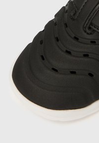 Zapatilla negra con parte superior texturizada en ondas, perforaciones y suela blanca. Cuenta con punta redondeada y acabado mate y suave.