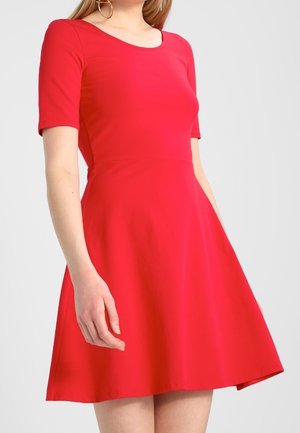 Kobieta ubrana w krótką, obcisłą czerwoną sukienkę z rozkloszowanym dołem, stojąca na tle jednolitego jasnoszarego tła.