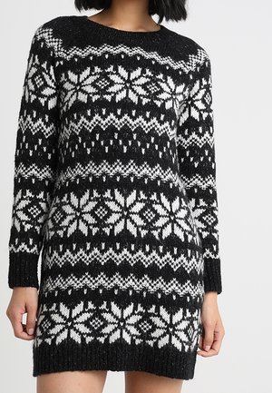Robe-pull à manches longues tricotée en noir et blanc avec des motifs de flocons de neige et géométriques, portée par une personne aux cheveux courts noirs.