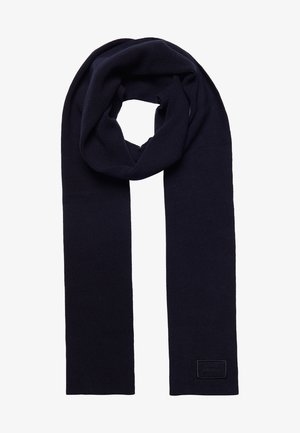 Écharpe en laine tricotée bleu marine foncé, pliée avec une petite étiquette en tissu rectangulaire cousue près d'une extrémité.