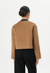 Bruine cropped jas met lange mouwen en een marineblauwe kraag, met een gestructureerde stof en een achterkantnaaddetail bij de nek. Zwarte broek is gedragen.