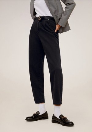 Personne portant un pantalon noir taille haute à jambes fuselées, des chaussettes blanches, des mocassins noirs avec détail chaîne dorée, une chemise blanche et un blazer à carreaux.