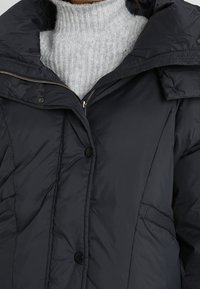 Tmavě modrá prošívaná bunda s vysokým límcem, zapínáním na zip vpředu a zapínáním na knoflíky. Má boční kapsy a prošívanou strukturu. Pod ní je měkký šedý svetr.