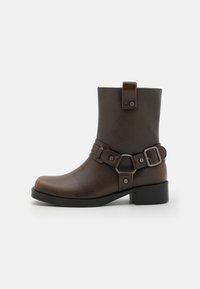 Bottine synthétique brune à semelle plate, avec un strap décoratif et un détail de boucle, texture lisse et design à bout arrondi.