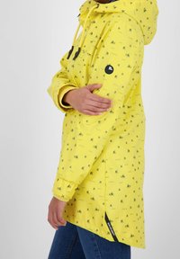 Gul regnjakke med mønster af små både, snørelukning i hætten og sorte detaljer på brystet og ærmet. Lange ærmer og sideslidser.