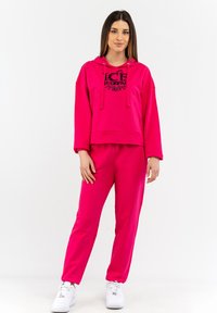 Felpa rosa con testo grafico nero, caratterizzata da una vestibilità comoda e cappuccio con cordini, abbinata a pantaloni joggers rosa coordinati. Sneakers bianche completano l'outfit.