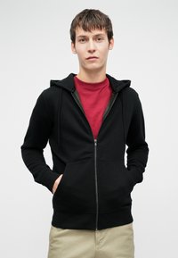 Mladý muž stojí s rukami v kapsách, oblečený v čiernej zipsovej mikine cez červené tričko a béžové nohavice, na pozadí s jednoduchou bielou farbou.