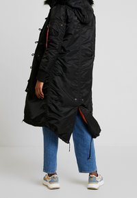 Czarna puchowa kurtka z kapturem wykończonym sztucznym futrem, bocznymi kieszeniami i detalami ze sznurkiem. Posiada rozcięcie z tyłu oraz kontrastową pomarańczową podszewkę.