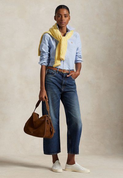 Woman wearing light blue button-up shirt, yellow sweater draped on shoulders, cropped jeans, white sneakers, and holding brown handbag.