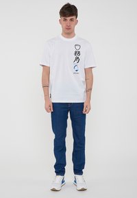 T-shirt bianco in cotone con grafiche nere e blu sul fronte, tra cui kanji e la scritta "KYOTO." Abbinato a jeans blu e sneakers bianche.