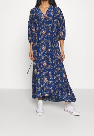 Woman wearing a blue floral long dress with 3/4 sleeves and white high-top sneakers, standing against a plain white background.