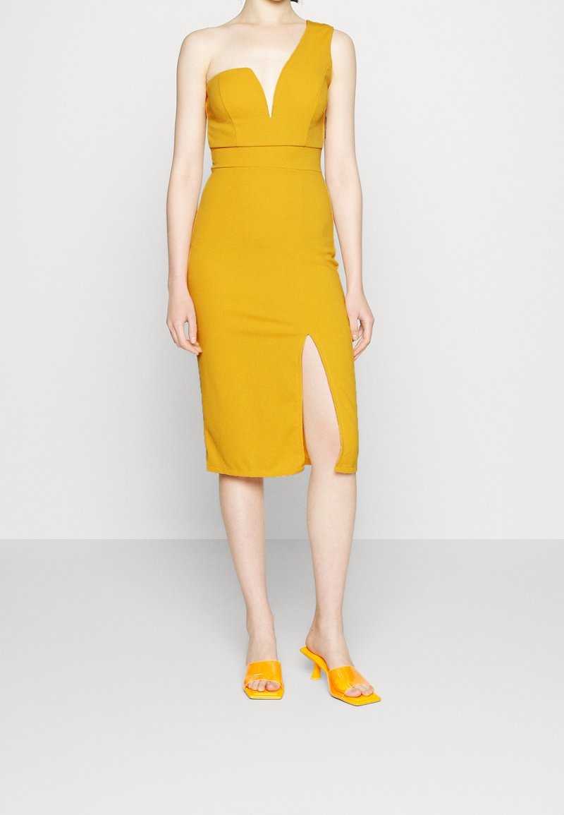 Femme portant une robe midi jaune moutarde asymétrique avec une fente à l'avant et des sandales à talons jaunes assorties, debout sur un fond clair.