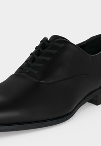 Chaussure habillée formelle en cuir noir avec une finition lisse et un laçage fermé sur fond blanc.