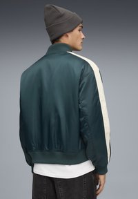 Satin bomberjack in donker teal met een crèmekleurige streep; heeft een geribbelde kraag, manchetten en zoom; gedragen over een wit shirt.