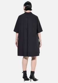 Abito nero a maniche corte con design strutturato, dettaglio di pieghe sul retro e silhouette dritta, abbinato a stivali neri con tacco alto.