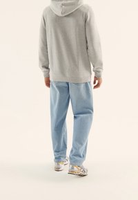 Persona con sudadera con capucha gris, vaqueros holgados azul claro y zapatillas blancas con detalles dorados, vista desde atrás sobre un fondo liso.