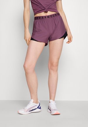 Kobieta nosząca fioletowe krótkie spodenki do biegania Under Armour i pasujący top z supłem, stojąca na szarej podłodze w białych i fioletowych butach sportowych.