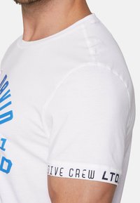 Bílá bavlněná tričko s modrým potiskem na přední straně a štítkem na rukávu s nápisem "DIVE CREW LTD." Přiléhavý střih, krátké rukávy.