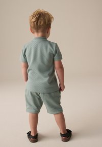 Light green short-sleeve polo shirt with textured pattern, paired with matching shorts. Child's black sandals with buckle detail visible.
